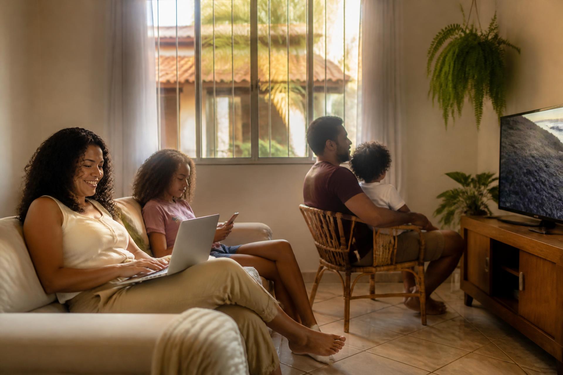 Família brasileira em casa: mãe no notebook, filha no celular e pai com a criança assistindo à TV, no fim da tarde